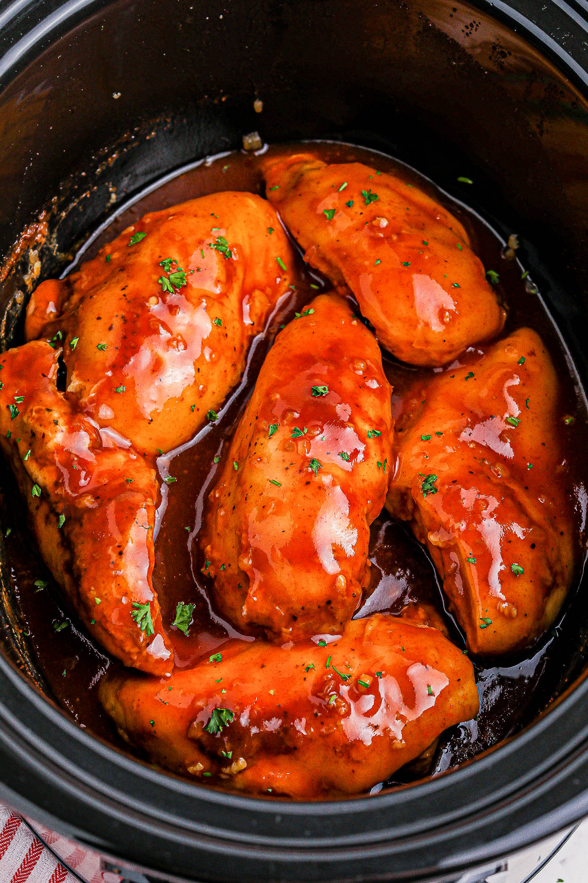 Slow cooker filled with five pieces of chicken breast coated in a glossy barbecue sauce, garnished with chopped parsley.