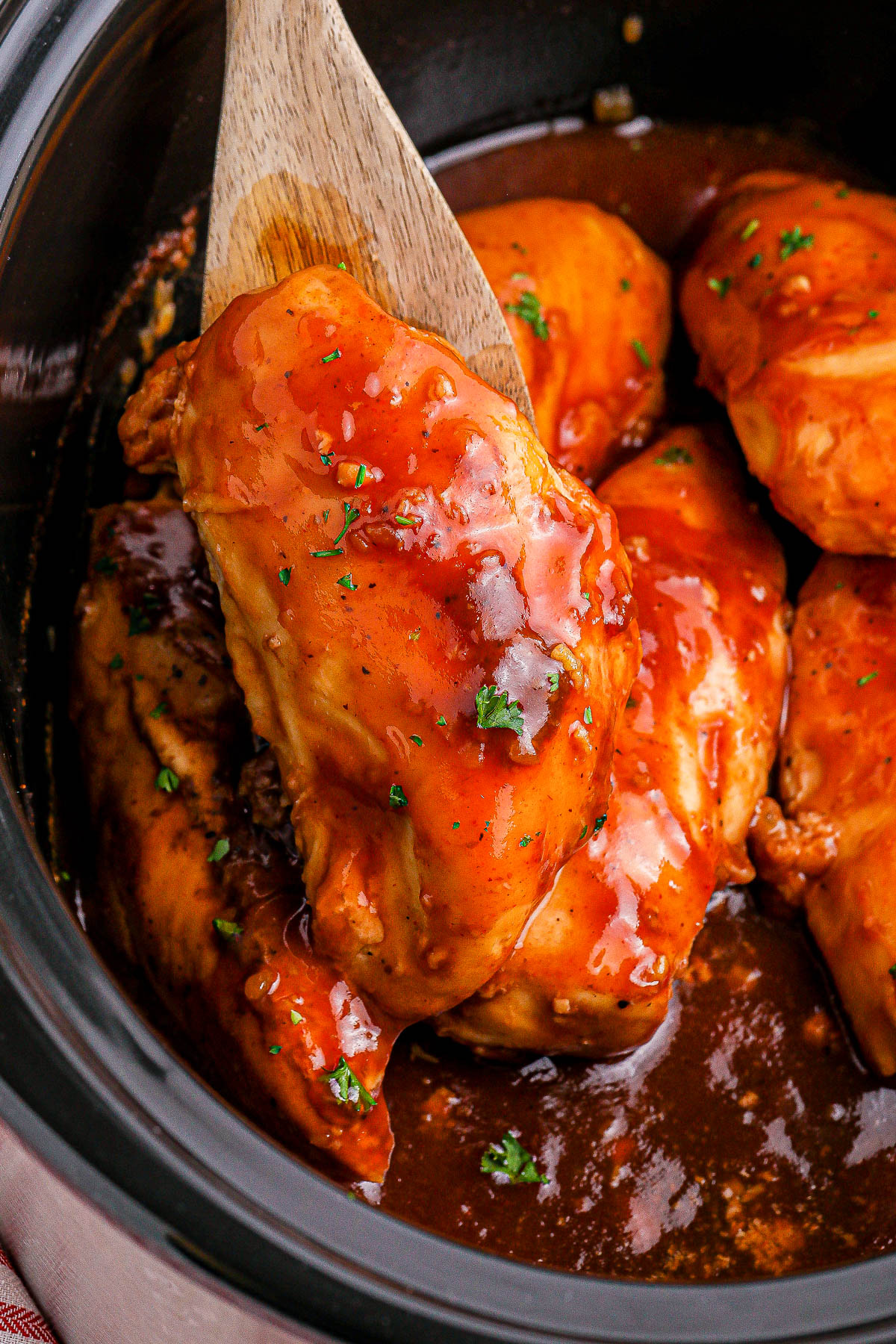 A wooden spoon lifts a cooked chicken breast covered in a glossy BBQ sauce, with more chicken pieces and sauce visible in a pot.