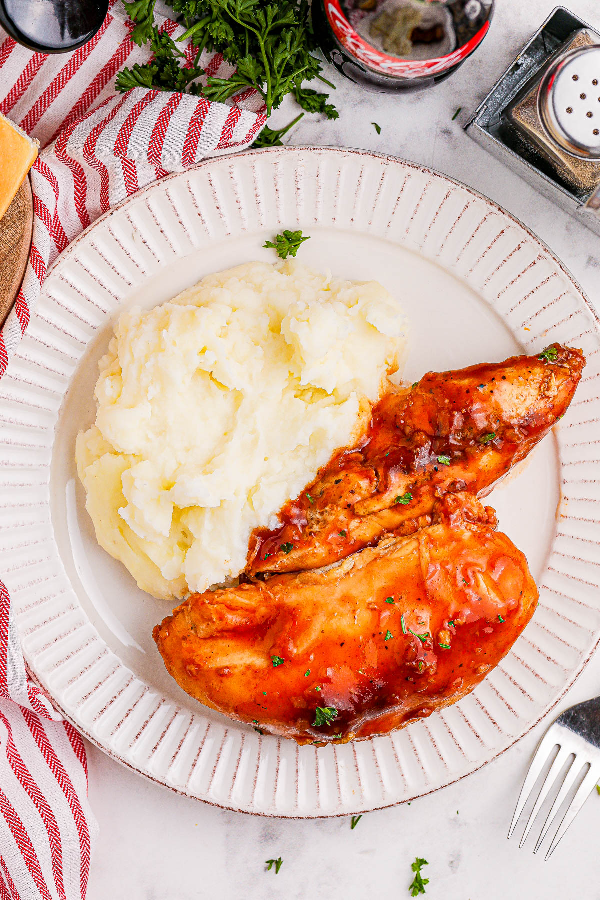 Plate with BBQ-glazed chicken and mashed potatoes, garnished with parsley. A fork, salt shaker, and soda bottle visible in the background.