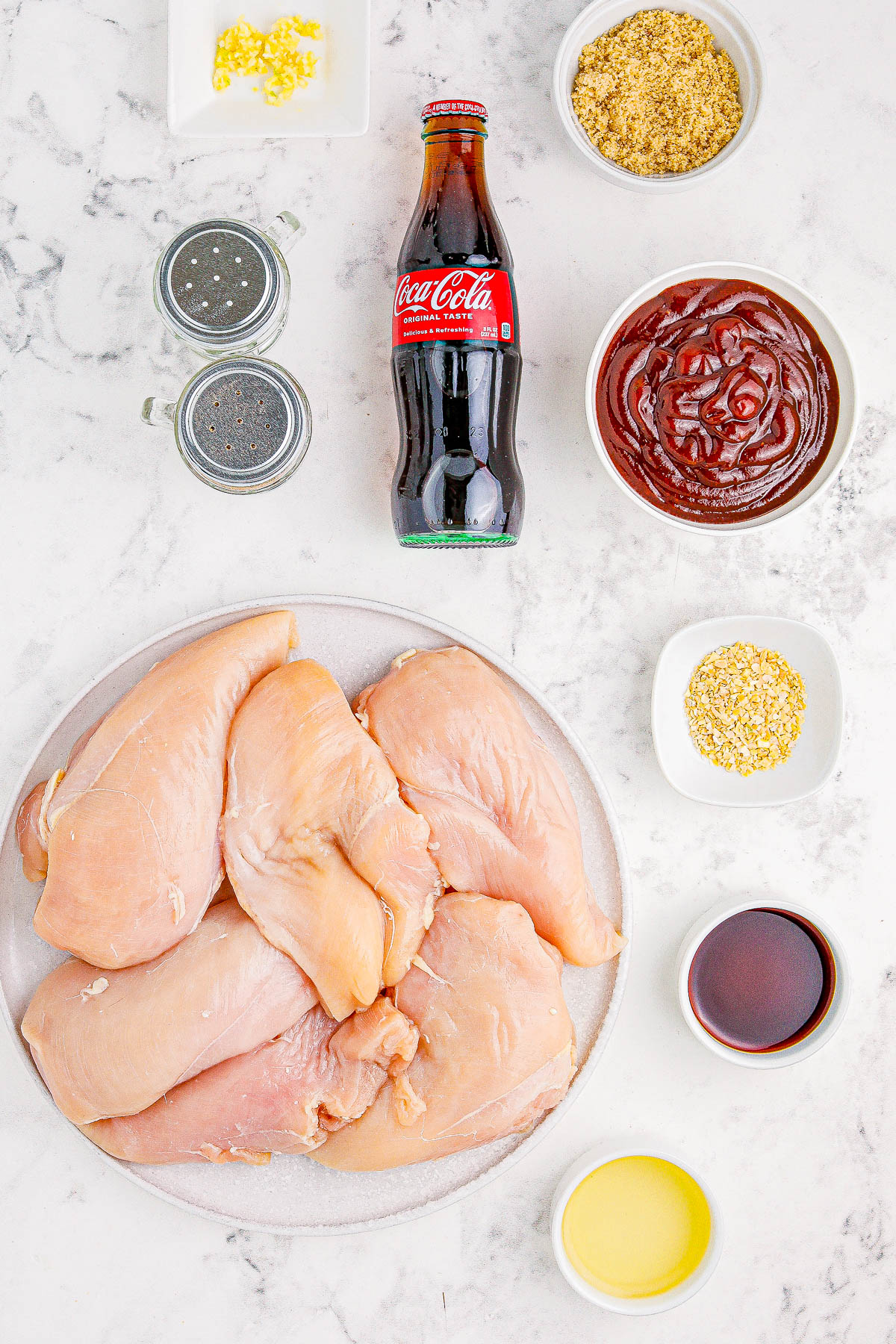 A variety of ingredients including raw chicken breasts, a bottle of Coca-Cola, bowls containing minced garlic, breadcrumbs, marinade sauce, and other seasonings, are displayed on a marble surface.