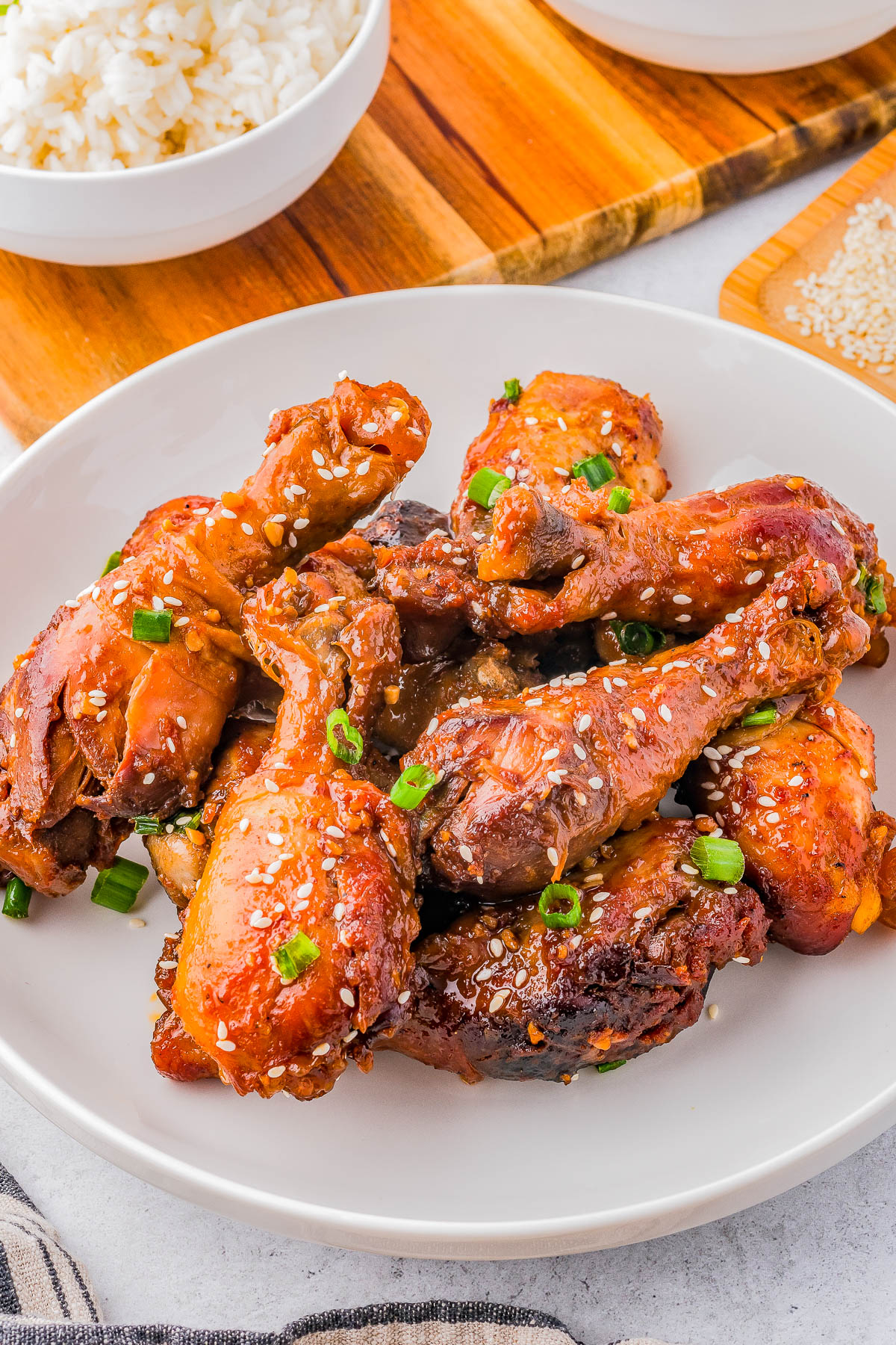 Plate of glazed chicken drumsticks garnished with green onions and sesame seeds, placed on a wooden board next to a bowl of white rice.