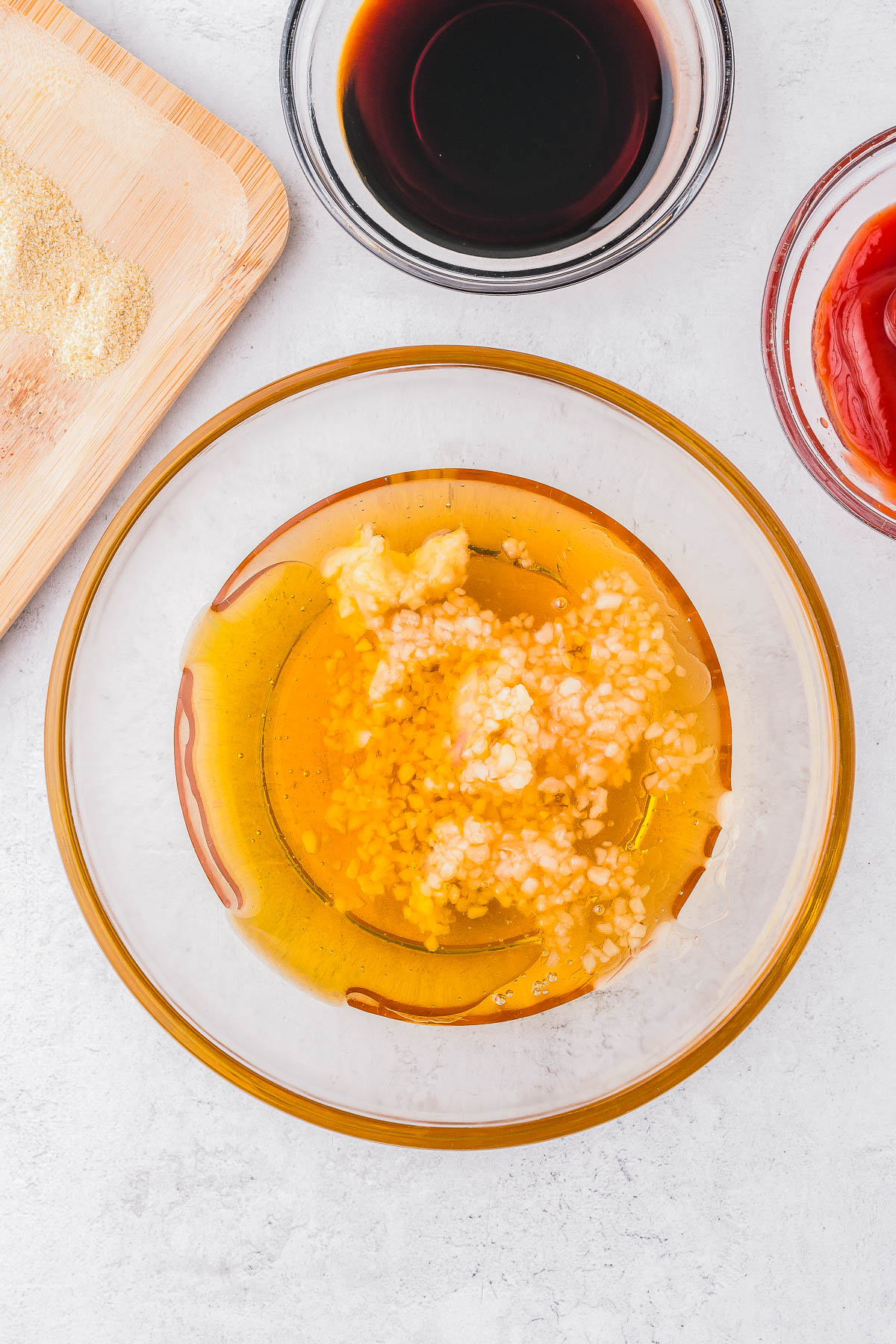 A bowl containing honey, minced garlic, and ginger. Surrounding the bowl are small dishes with ketchup and dark-colored sauce, and a wooden board with breadcrumbs.