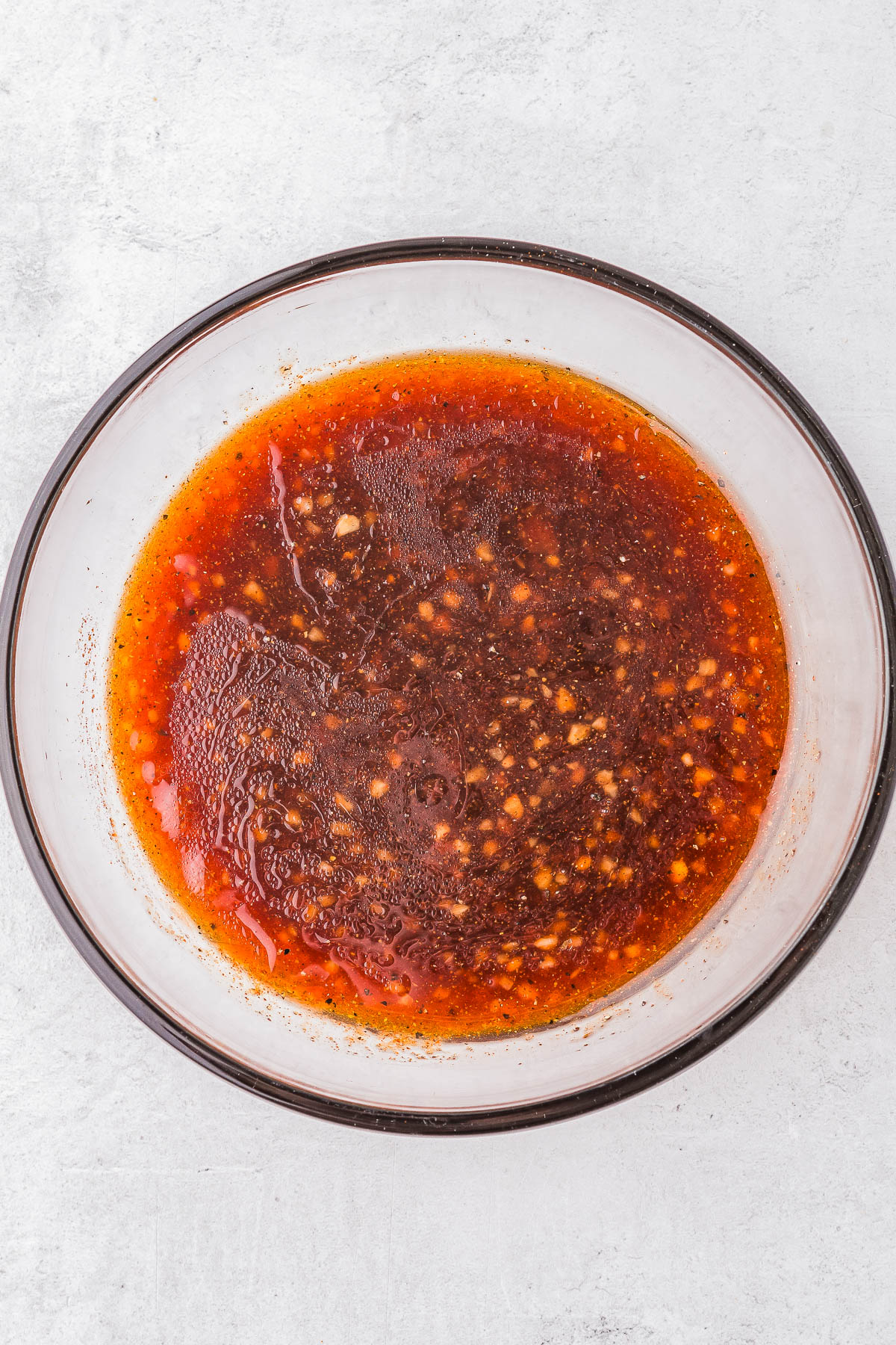 A glass bowl containing a reddish-brown liquid mixture with visible garlic and seasoning particles.