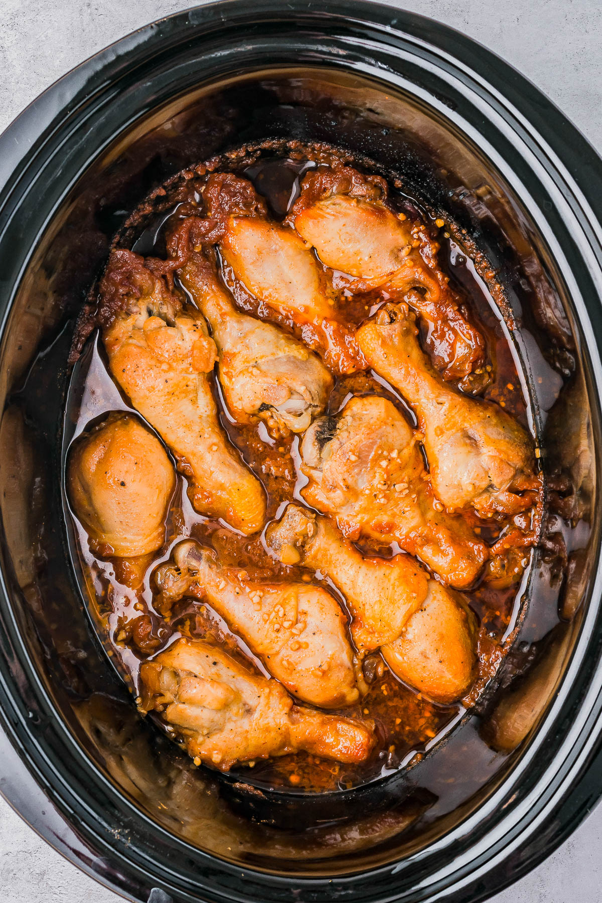 Slow cooker filled with cooked chicken drumsticks in a thick, reddish-brown sauce.