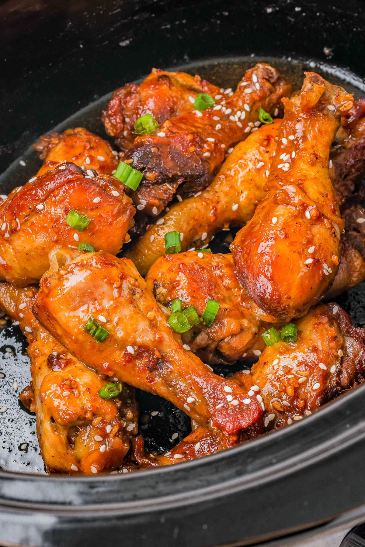 Close-up of glazed chicken drumsticks garnished with sesame seeds and chopped green onions in a black dish.