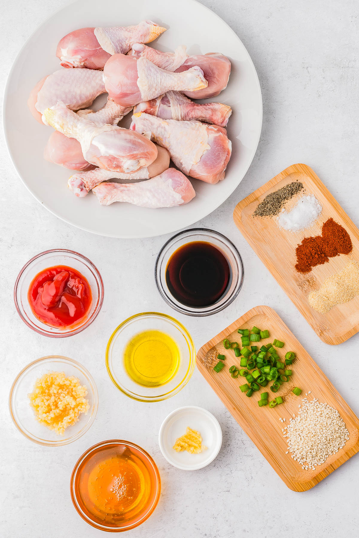 A plate of raw chicken drumsticks, bowls of various sauces and oils, chopped green peppers, minced garlic, and spices arranged on a white surface.