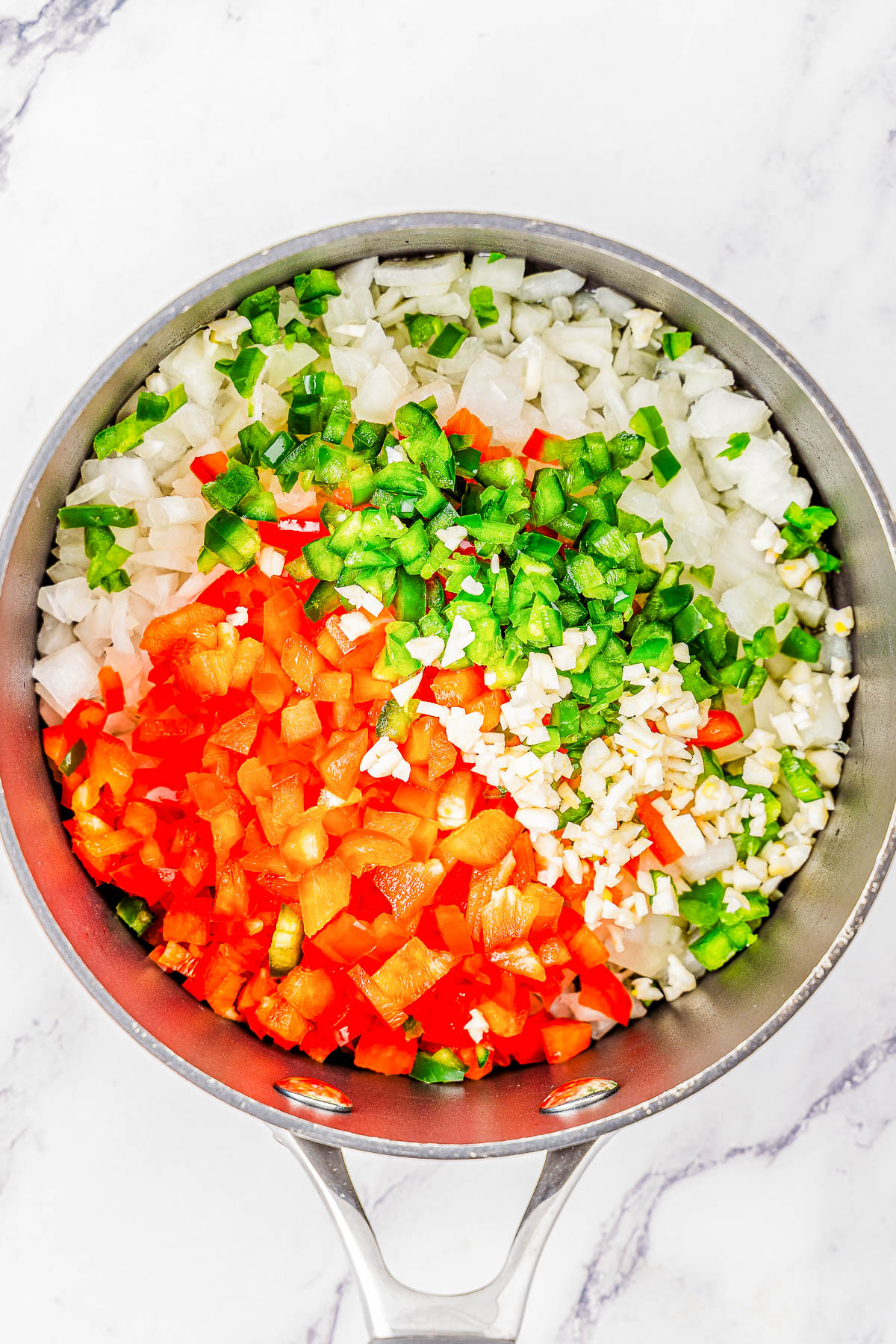 A pot filled with chopped red bell peppers, green bell peppers, onions, and garlic on a white marble surface.