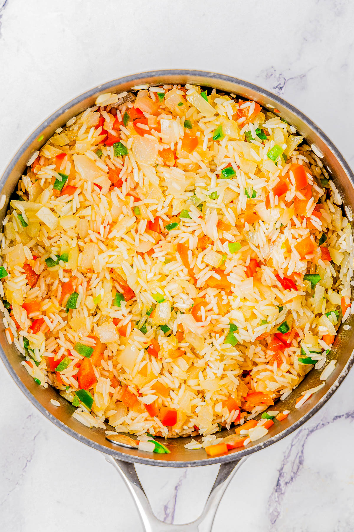 A stainless steel pot filled with cooked rice mixed with diced vegetables including red bell peppers, green onions, and possibly carrots, resting on a white marble surface.