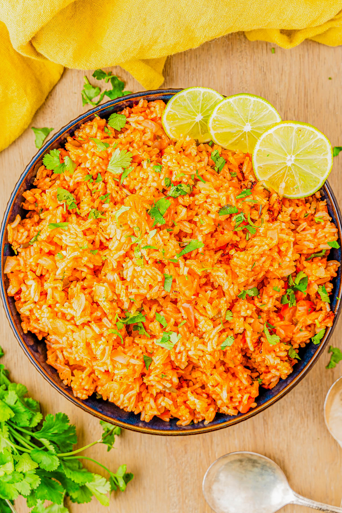 A bowl of Mexican rice garnished with fresh cilantro and lime slices, placed on a wooden surface with a yellow cloth and cilantro sprigs nearby.