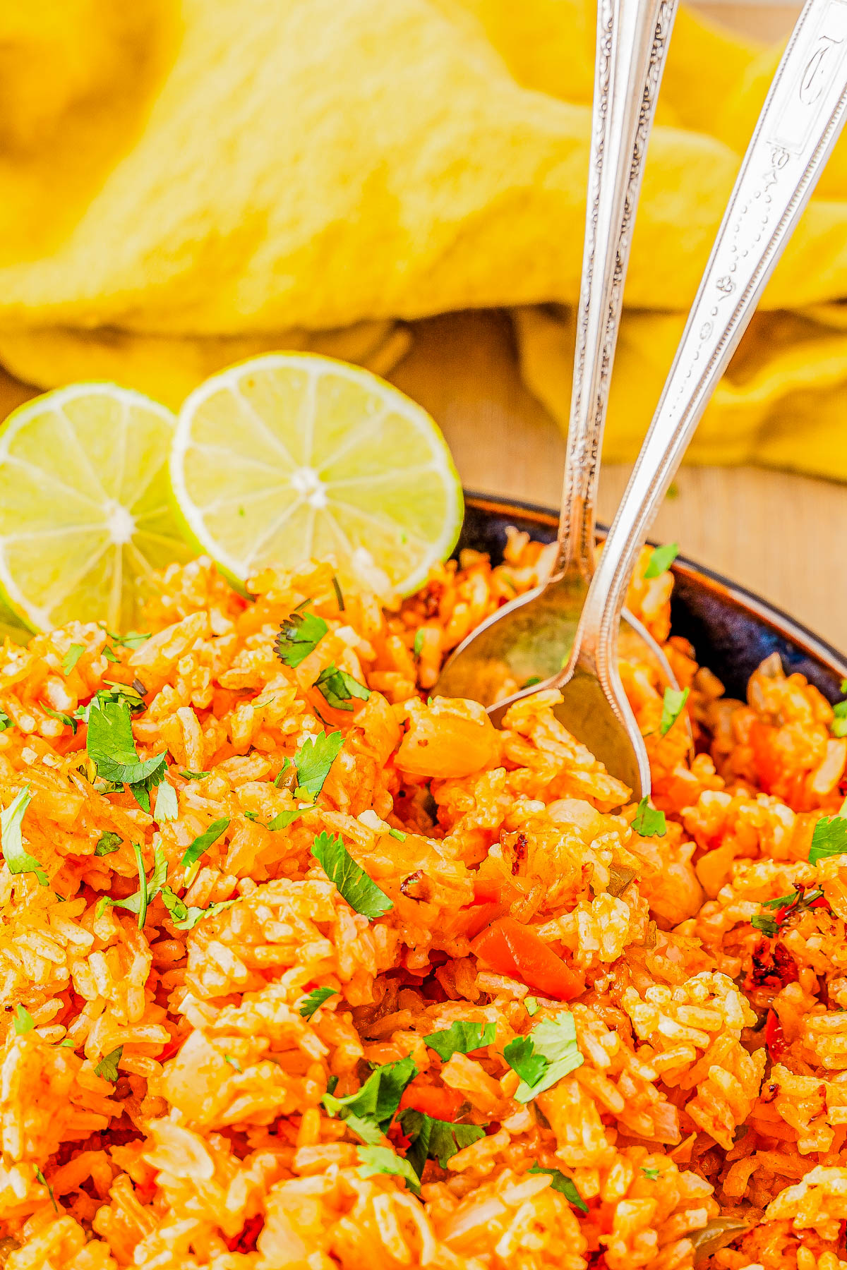 Close-up of a plate of seasoned rice with chopped vegetables, garnished with cilantro. Two slices of lime and two silver forks are also on the plate. A yellow cloth is in the background.