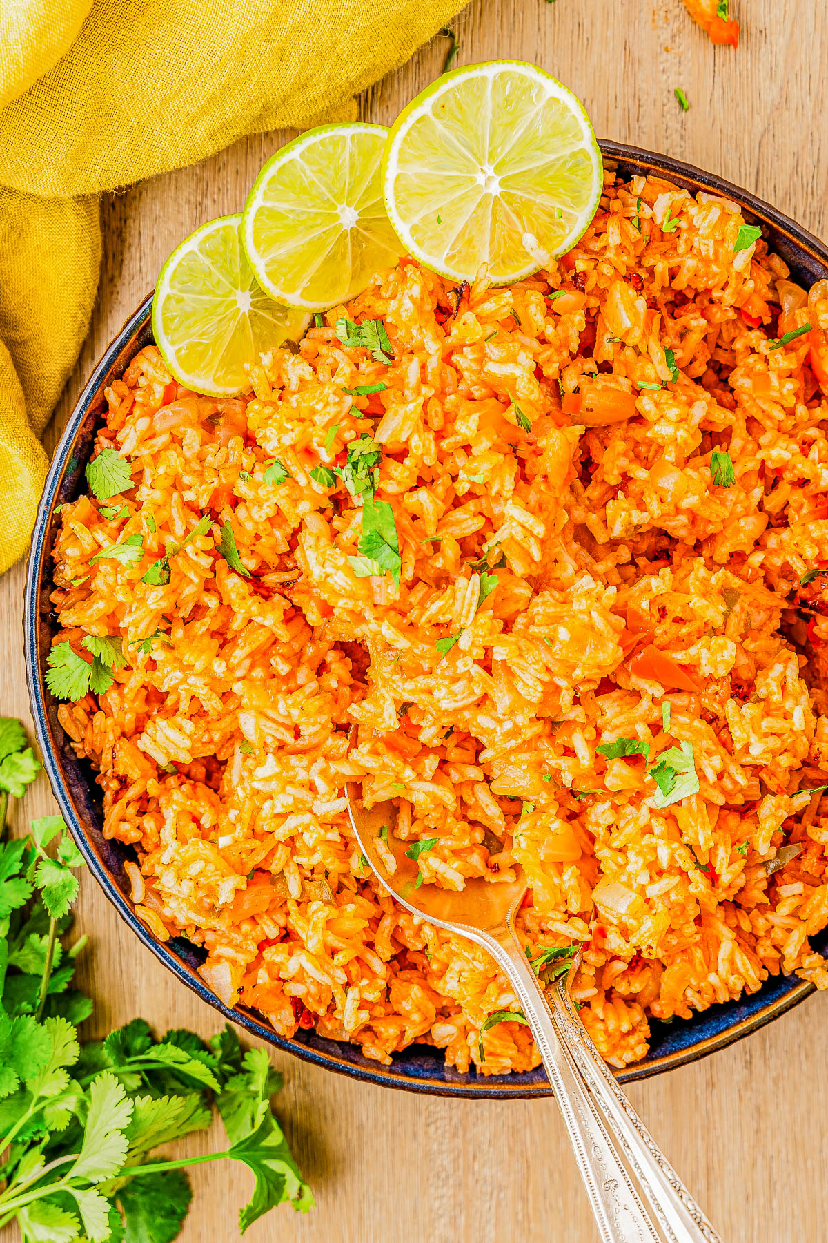 A bowl of seasoned rice garnished with three lime slices and cilantro, placed on a wooden surface with a yellow cloth and cilantro leaves nearby.