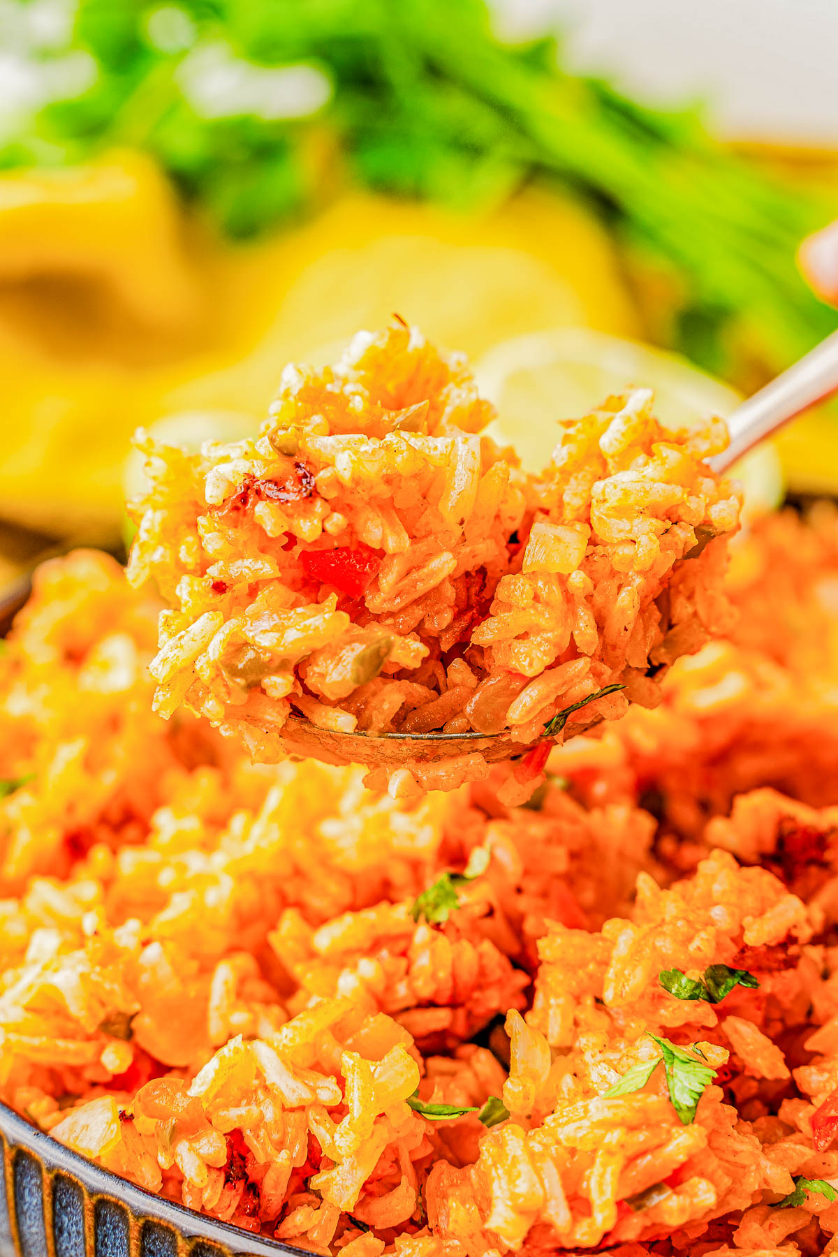 Close-up of a spoon holding a serving of cooked rice with herbs and vegetables, with more rice on the plate below and fresh greens in the background.