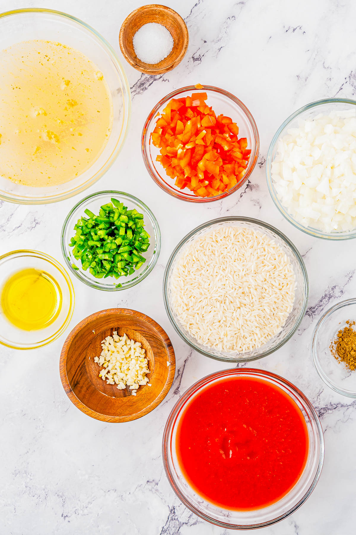 An arrangement of ingredients in bowls on a marble surface, including rice, diced tomatoes, diced onions, chopped green peppers, minced garlic, tomato sauce, oil, broth, and seasoning.