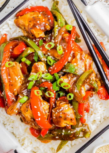 Colorful stir fry with sliced chicken, red and green bell peppers, topped with sesame seeds and green onions on a bed of white rice, accompanied by chopsticks.