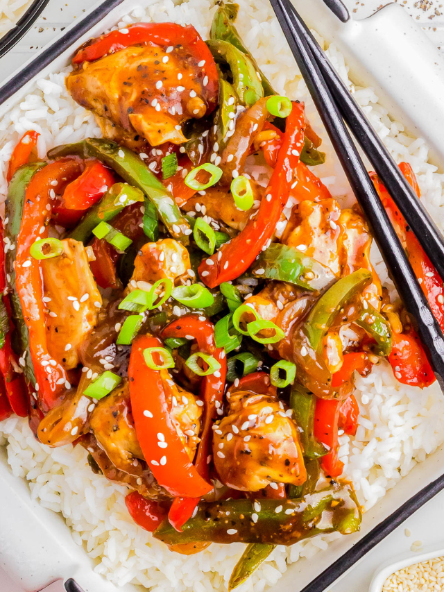 Colorful stir fry with sliced chicken, red and green bell peppers, topped with sesame seeds and green onions on a bed of white rice, accompanied by chopsticks.