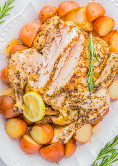 Roast chicken with sliced breast, garnished with rosemary and a lemon slice, surrounded by roasted potatoes on a white platter.