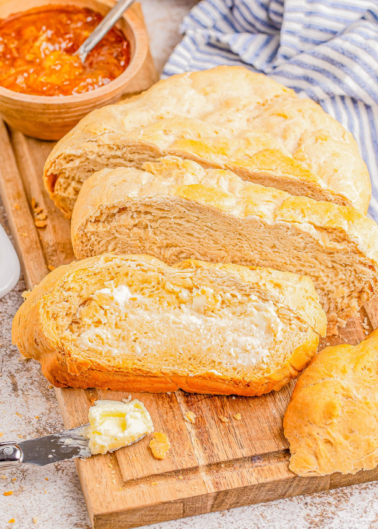 Sliced loaf of bread on a wooden board with a small bowl of orange marmalade and a blue-striped cloth in the background. Butter is being spread on one piece.