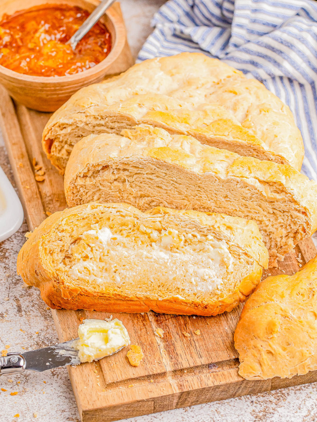 Sliced loaf of bread on a wooden board with a small bowl of orange marmalade and a blue-striped cloth in the background. Butter is being spread on one piece.