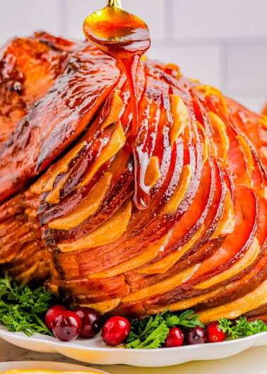 A glazed ham with sliced sections is displayed on a white plate, garnished with fresh cranberries and herbs, as honey is drizzled over the top.