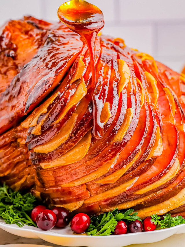 A glazed ham with sliced sections is displayed on a white plate, garnished with fresh cranberries and herbs, as honey is drizzled over the top.