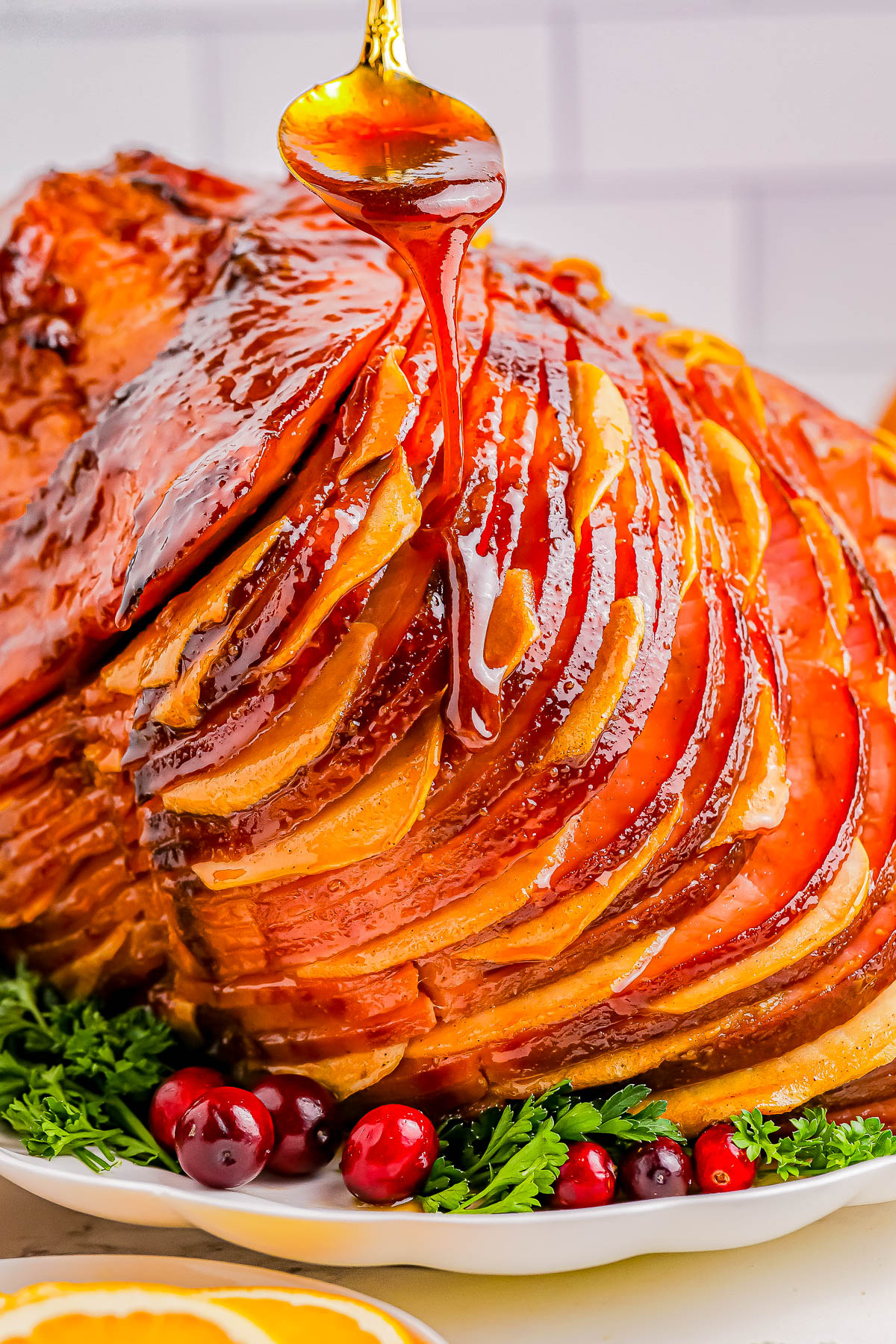 A glazed ham with sliced sections is displayed on a white plate, garnished with fresh cranberries and herbs, as honey is drizzled over the top.