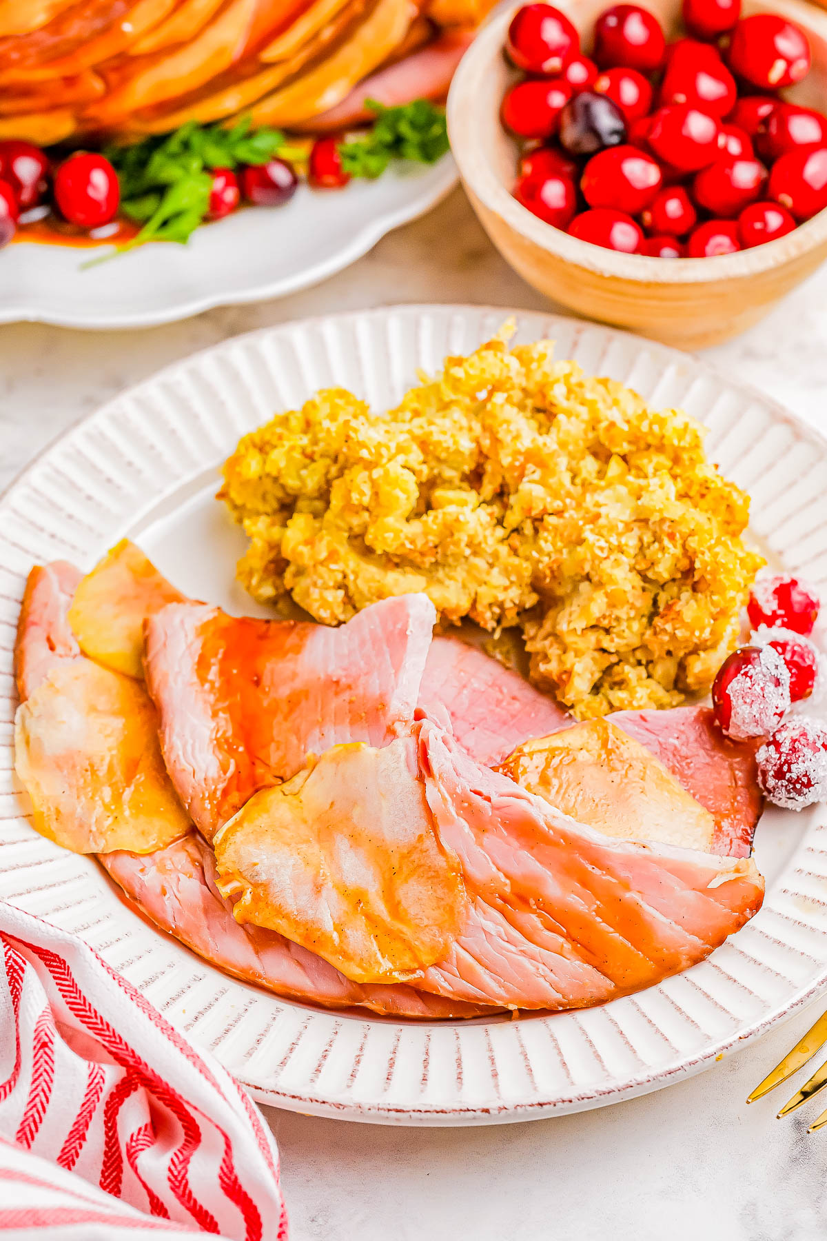 A plate with slices of ham, a serving of stuffing, and a few cranberries. A bowl of cranberries is in the background.