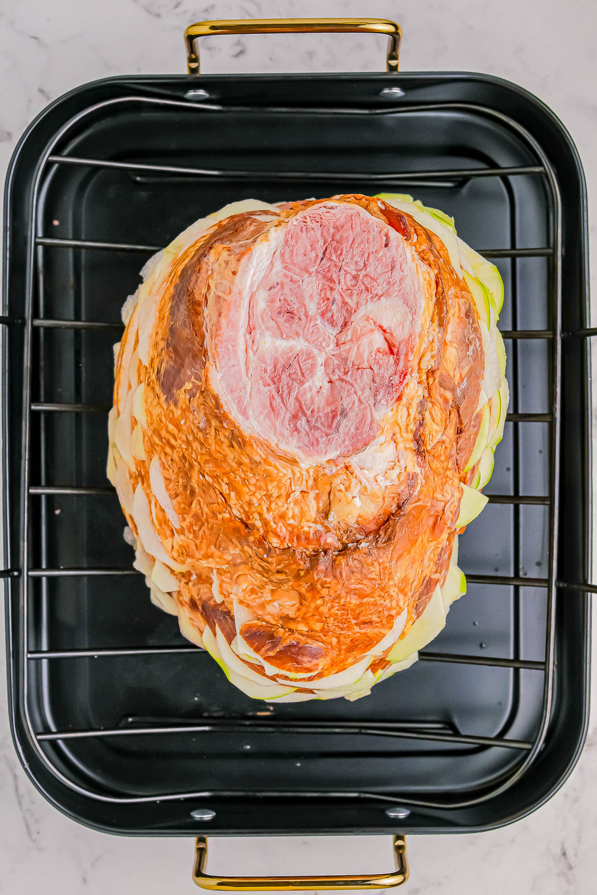 A raw ham sits on a bed of sliced onions in a black roasting pan with handles, ready for cooking.