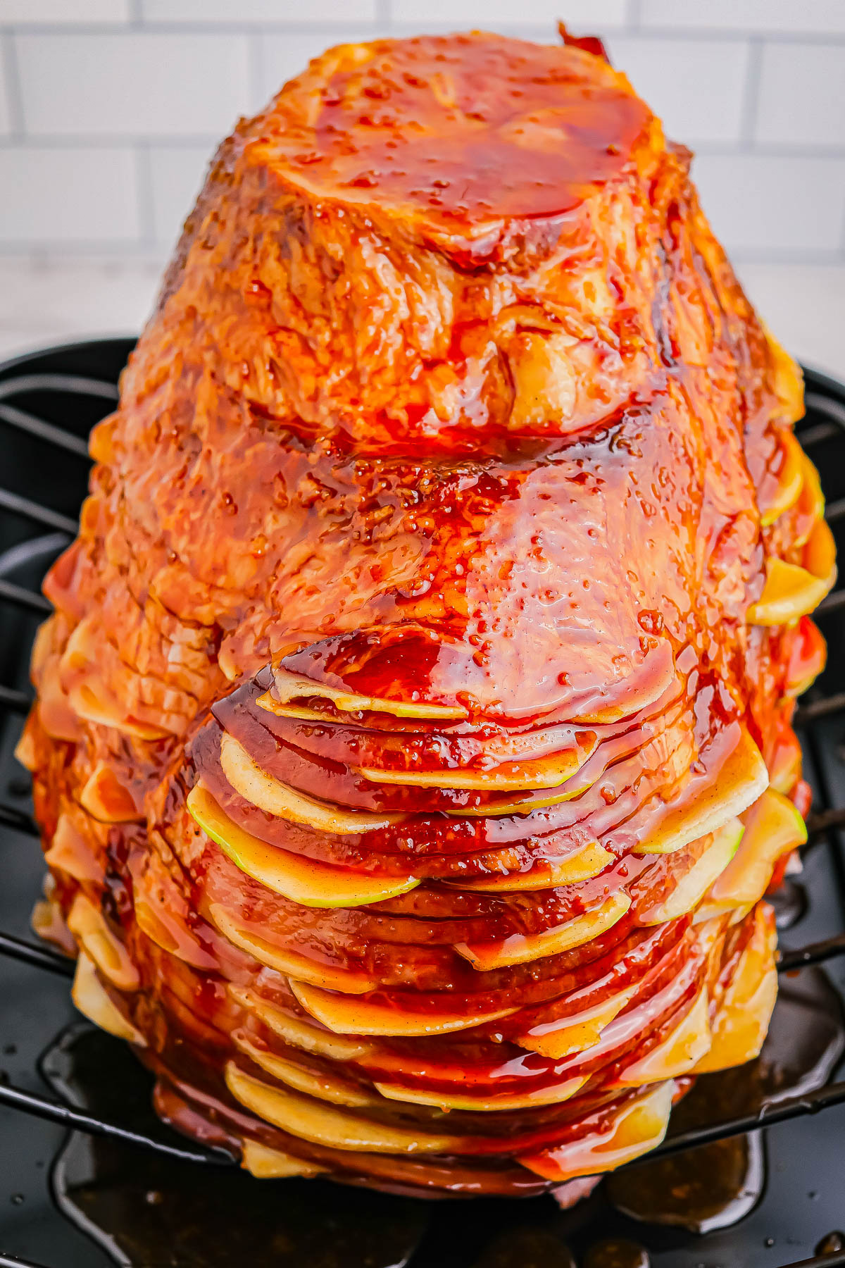 A glazed ham is shown on a roasting pan, covered in a shiny, caramel-colored sauce. The ham is spiral cut, revealing layers of meat.