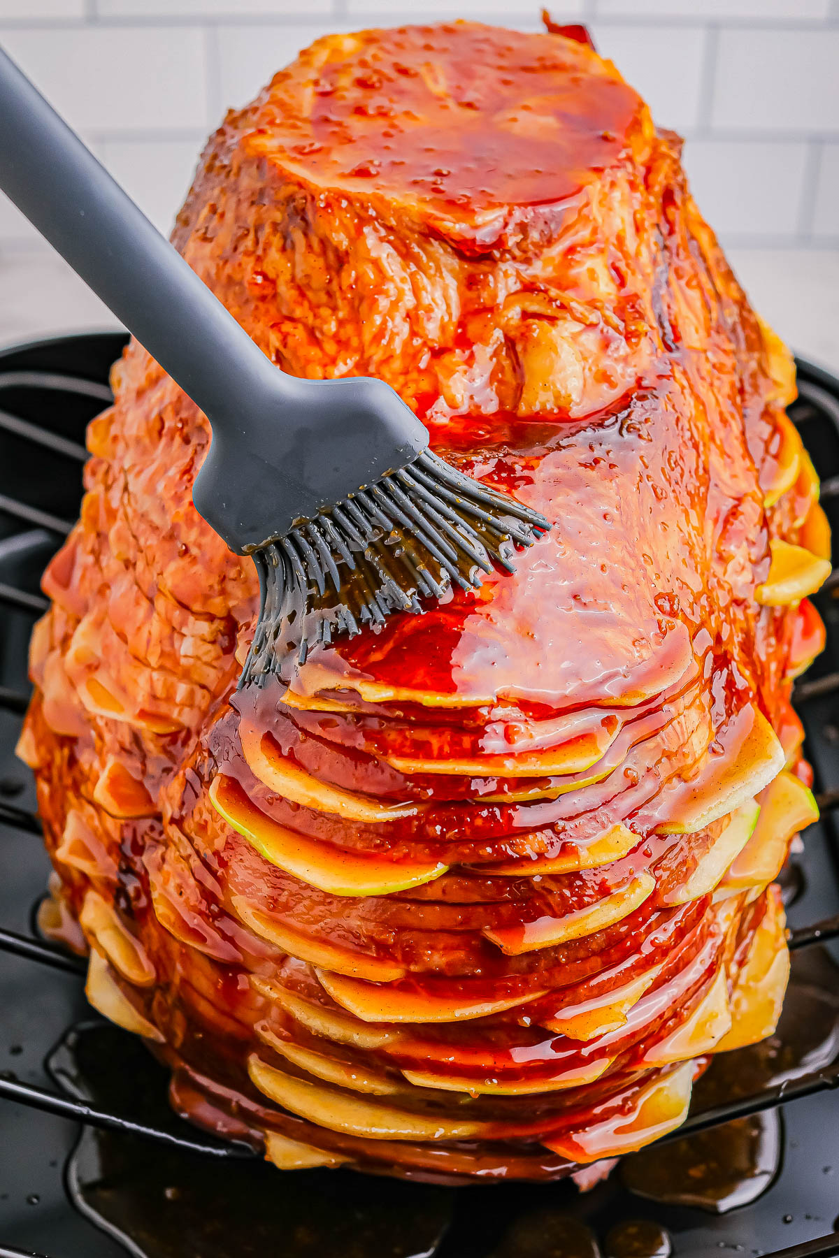 A layered stack of thinly sliced ham is being glazed with a brush, creating a shiny coating. The ham is resting on a black rack against a tiled background.