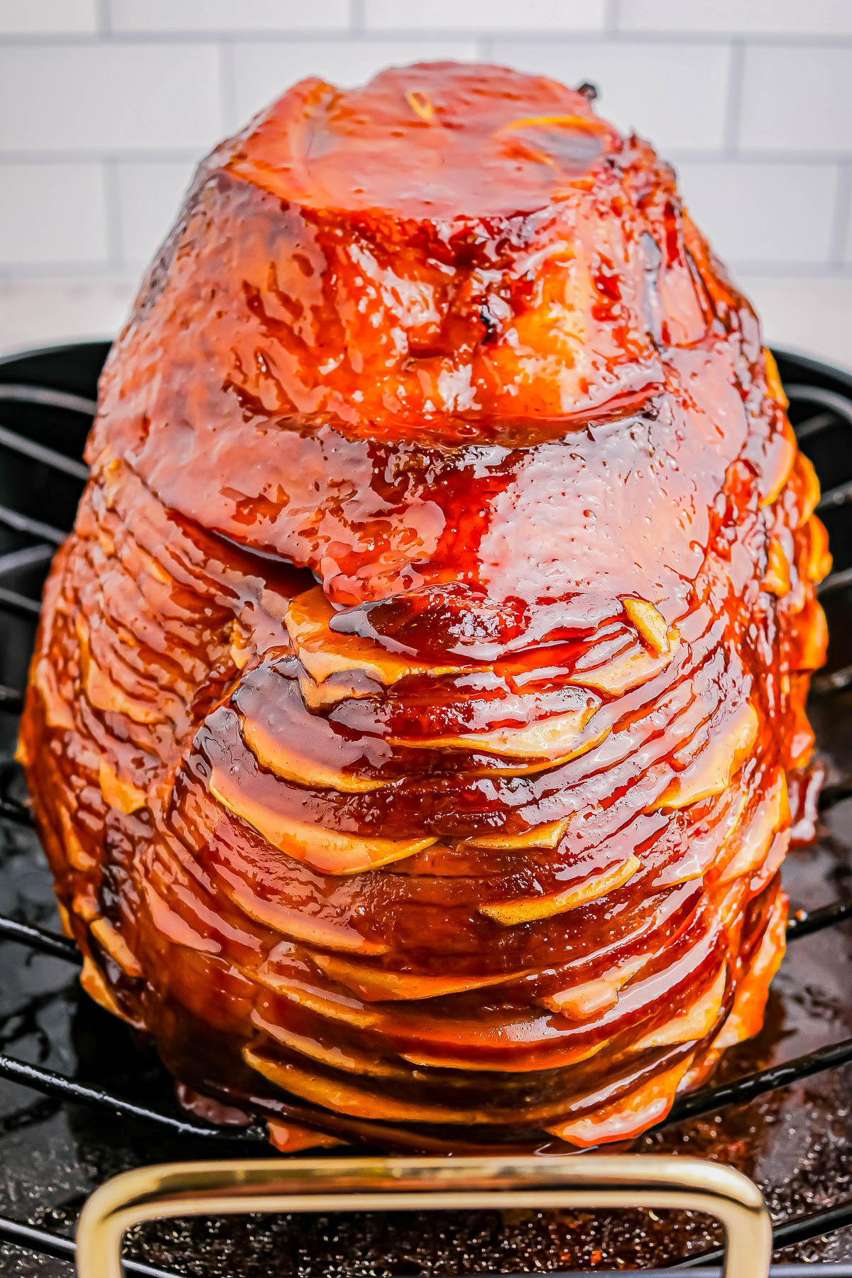 Glazed ham with spiral cuts placed on a rack in front of a white tiled background.