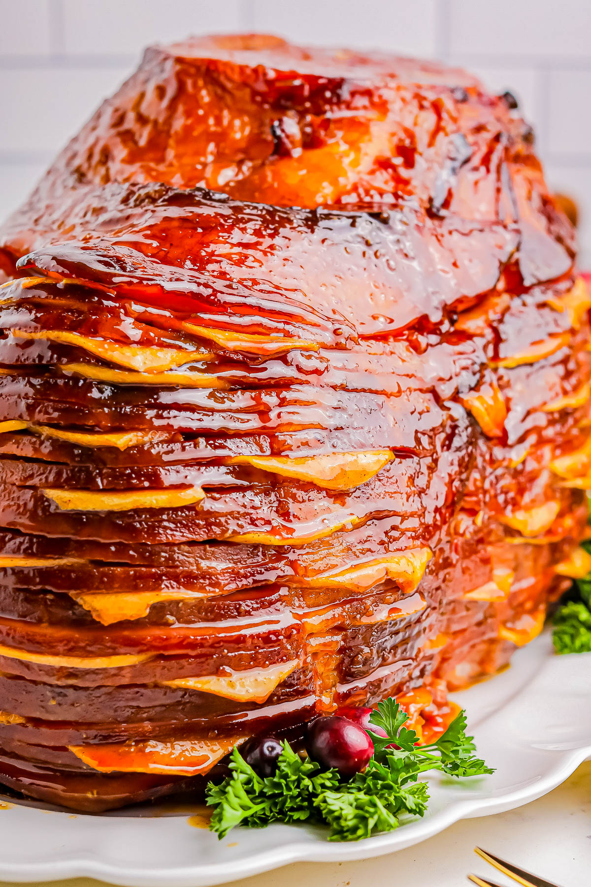 Close-up of a glazed spiral-cut ham on a white platter, garnished with sprigs of parsley and cranberries.