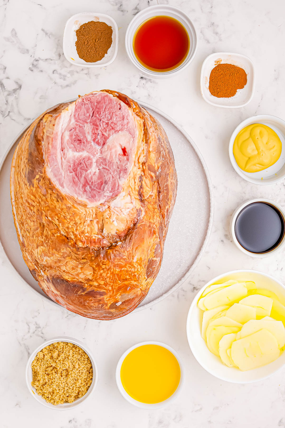 A whole ham surrounded by various ingredients, including sliced potatoes, mustard, and spices, on a marble surface.