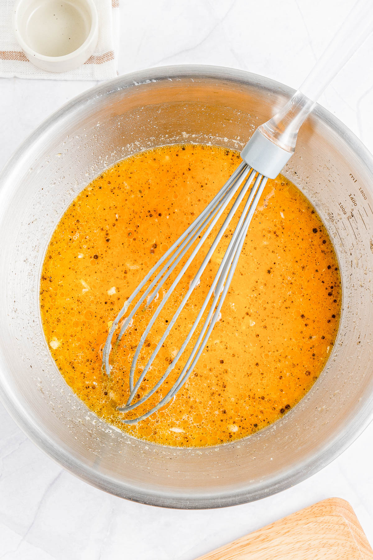 A mixing bowl contains an orange batter with visible spices and a whisk. A small empty bowl and a checked cloth are nearby.