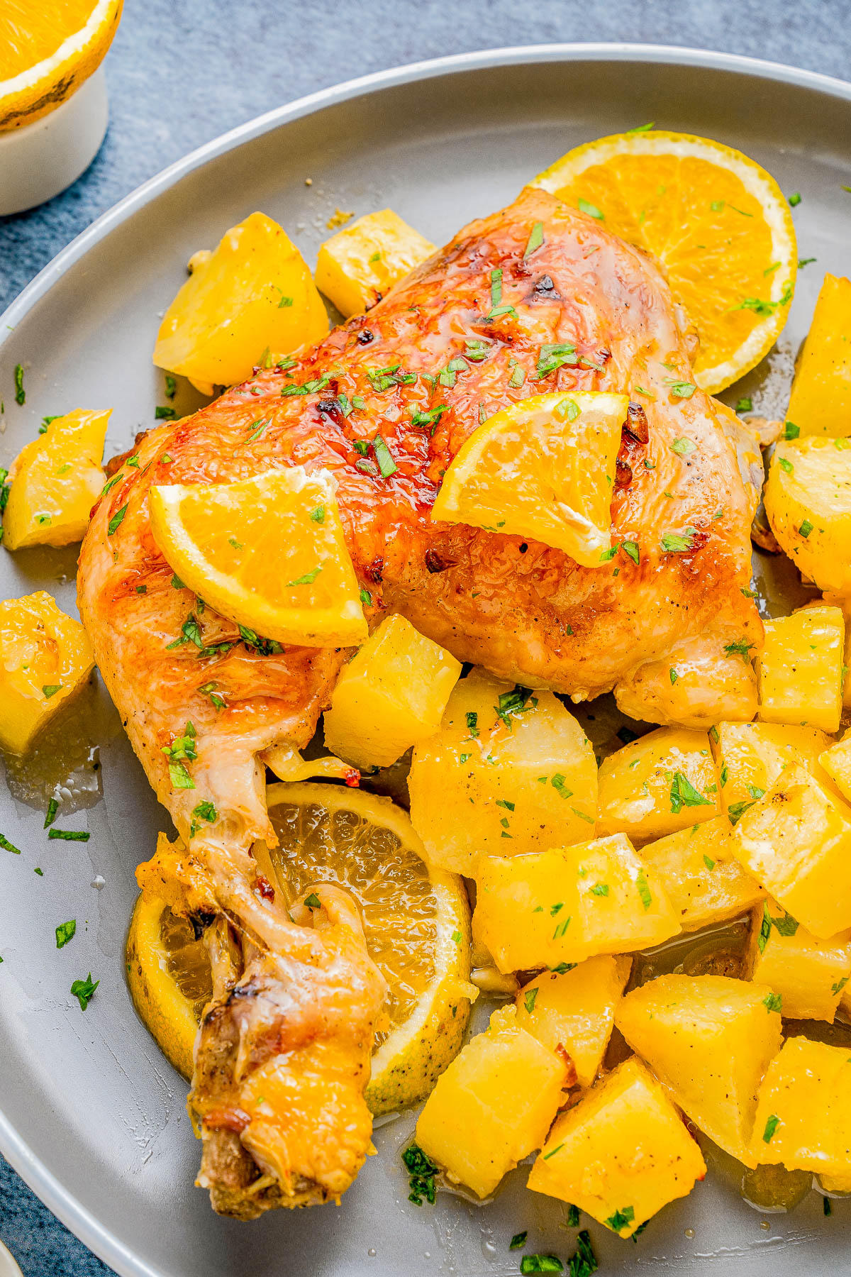 Roasted chicken leg with golden-brown skin is served with diced potatoes and orange slices, garnished with fresh herbs, on a gray plate.