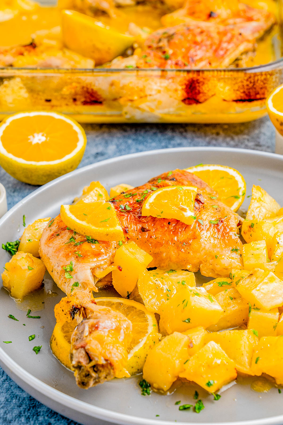 A plate of roasted chicken with potato chunks and lemon slices. A glass baking dish with more of the same dish is in the background.