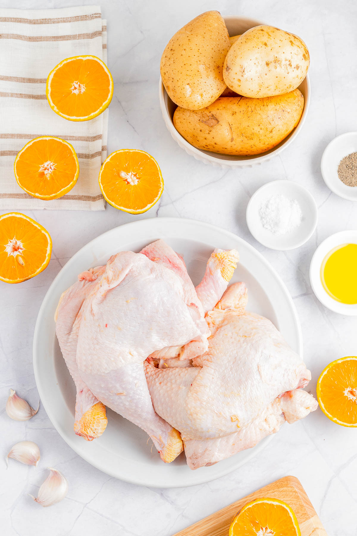 Two raw chickens on a white plate surrounded by halved oranges, whole potatoes in a bowl, garlic cloves, small bowls of salt, pepper, and oil on a marble surface with a striped cloth.