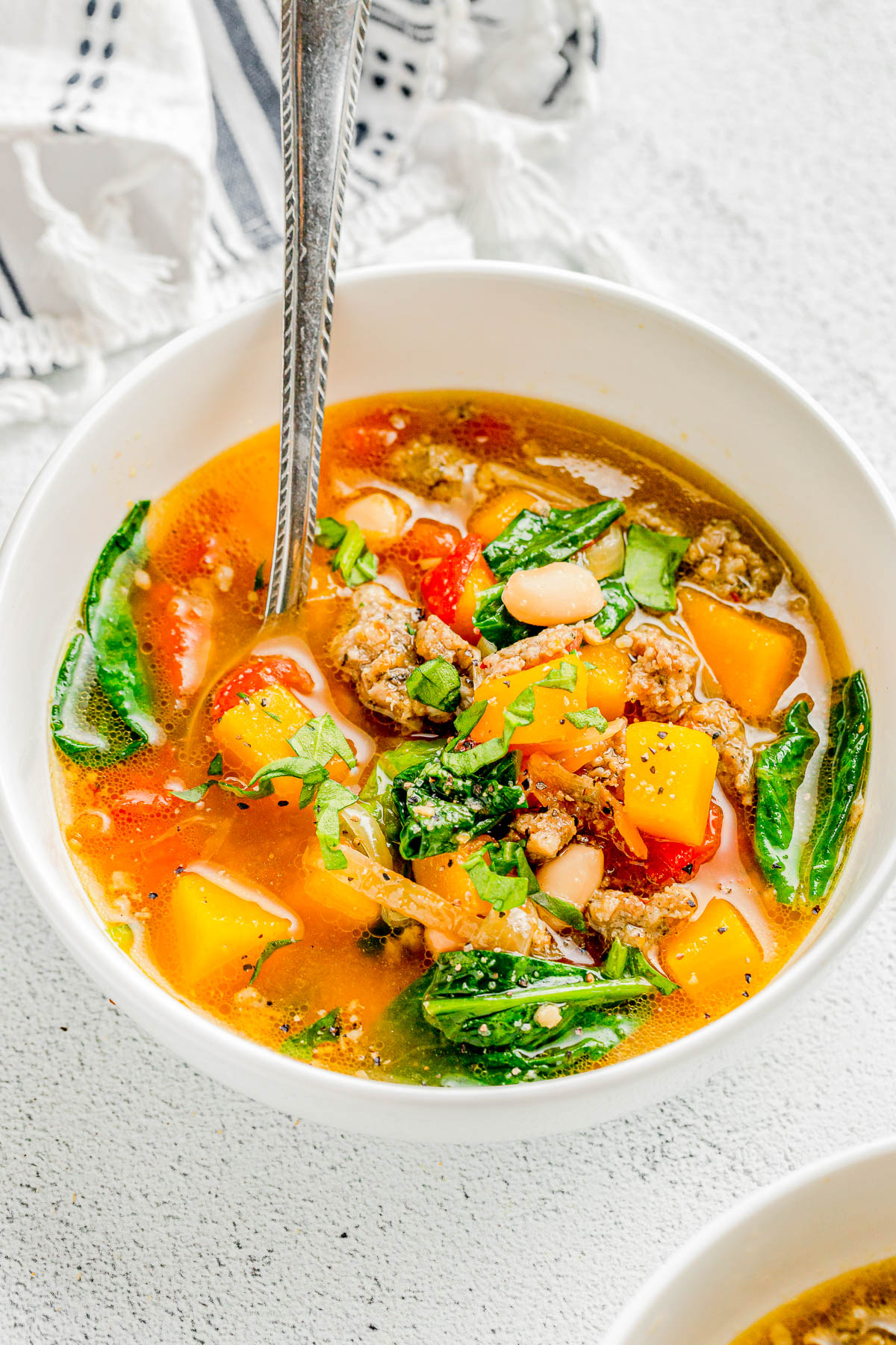 A bowl of soup with chunks of squash, greens, beans, and ground meat, garnished with herbs. A spoon stands upright in the bowl.