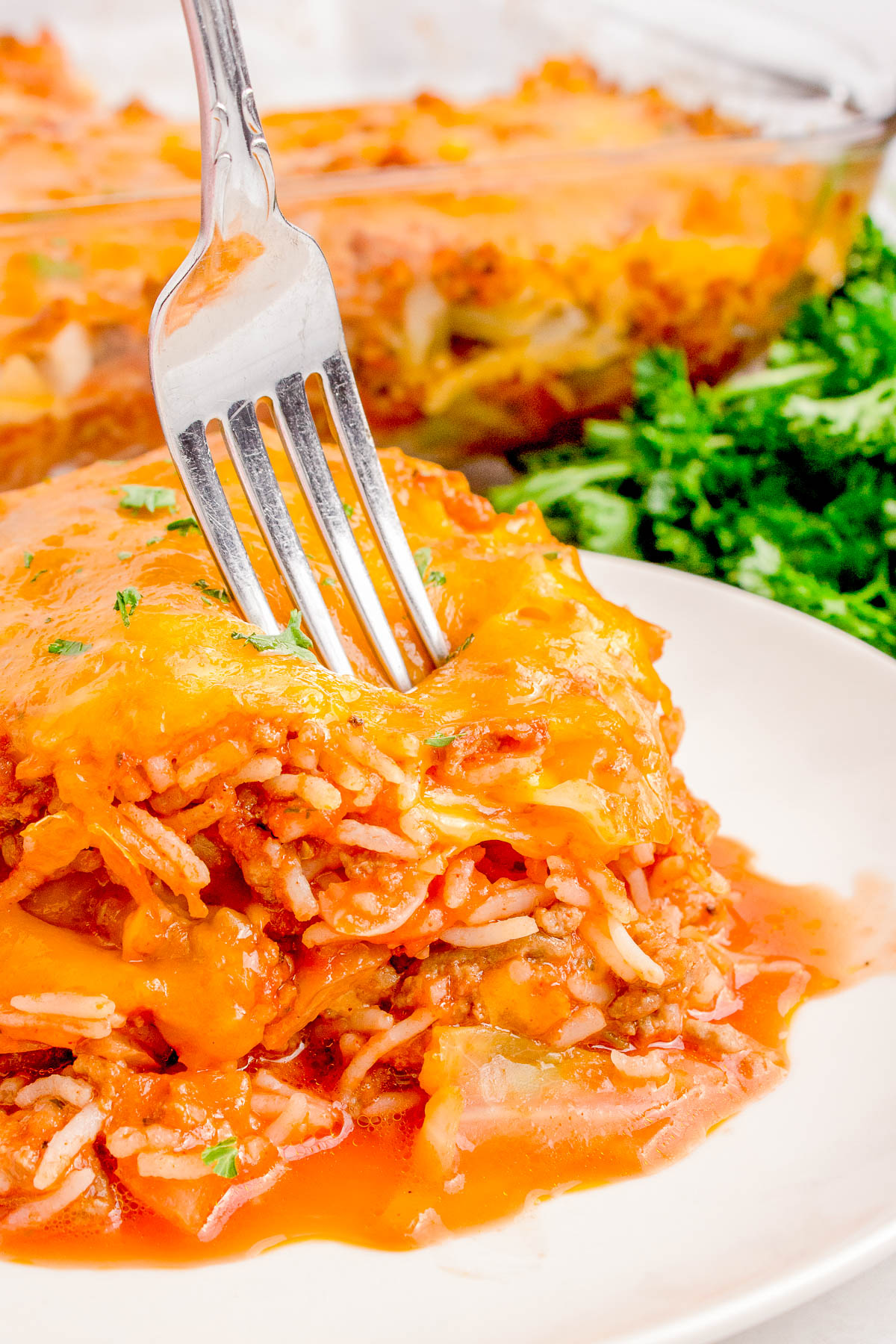 A fork resting on a slice of lasagna topped with melted cheese on a white plate, accompanied by a garnish of fresh parsley. A glass dish of lasagna is visible in the background.