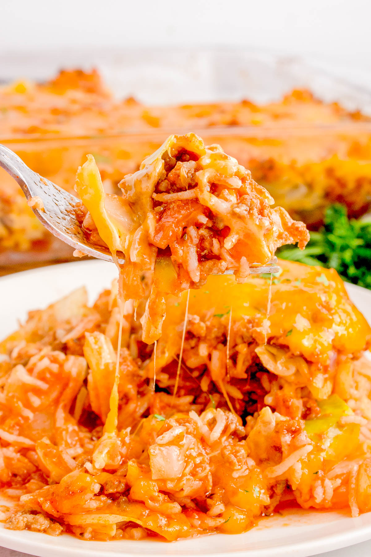 A fork lifts a cheesy spaghetti portion from a plate, revealing a casserole dish in the background.