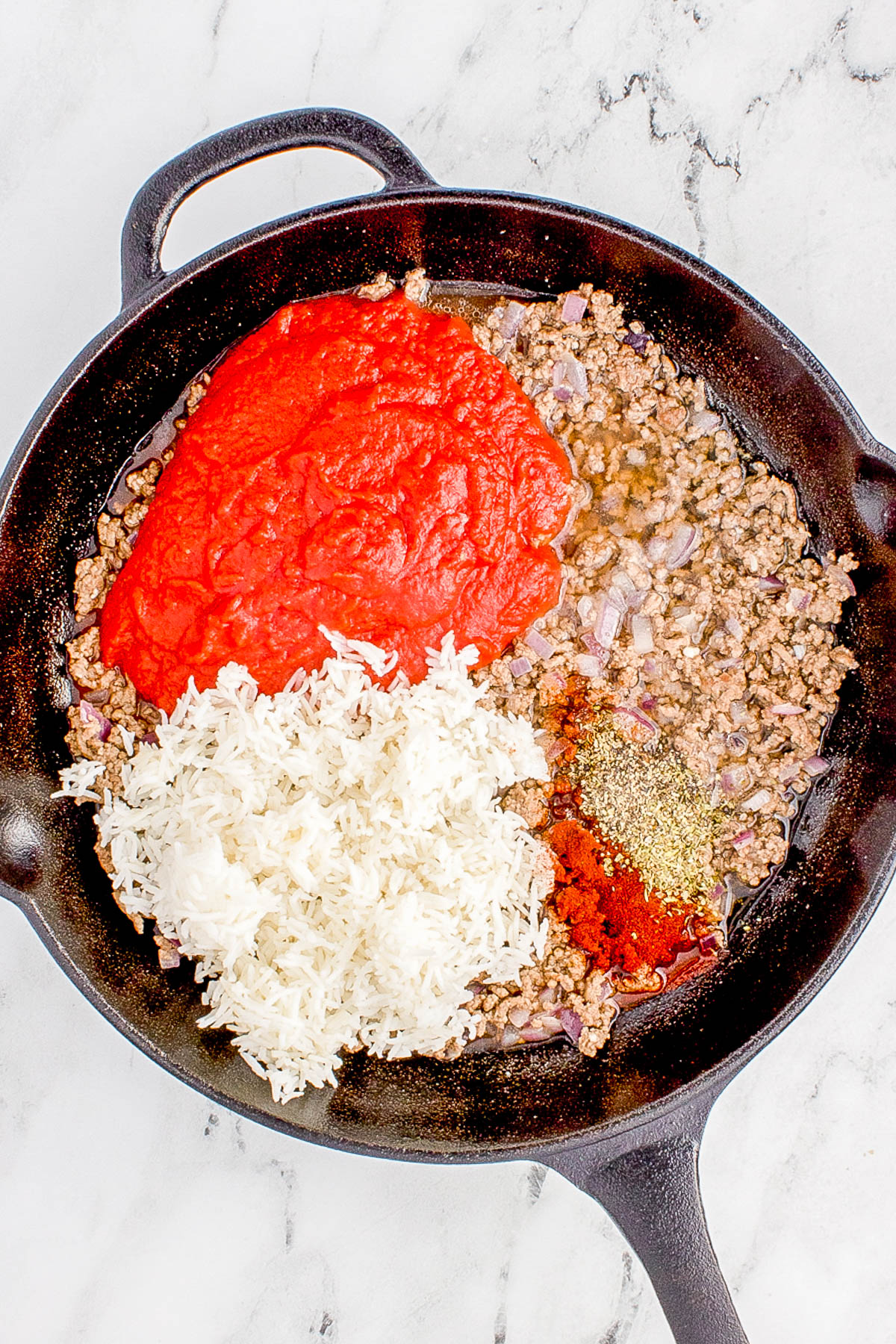 A skillet containing ground meat, tomato sauce, white rice, and various spices on a marble countertop.