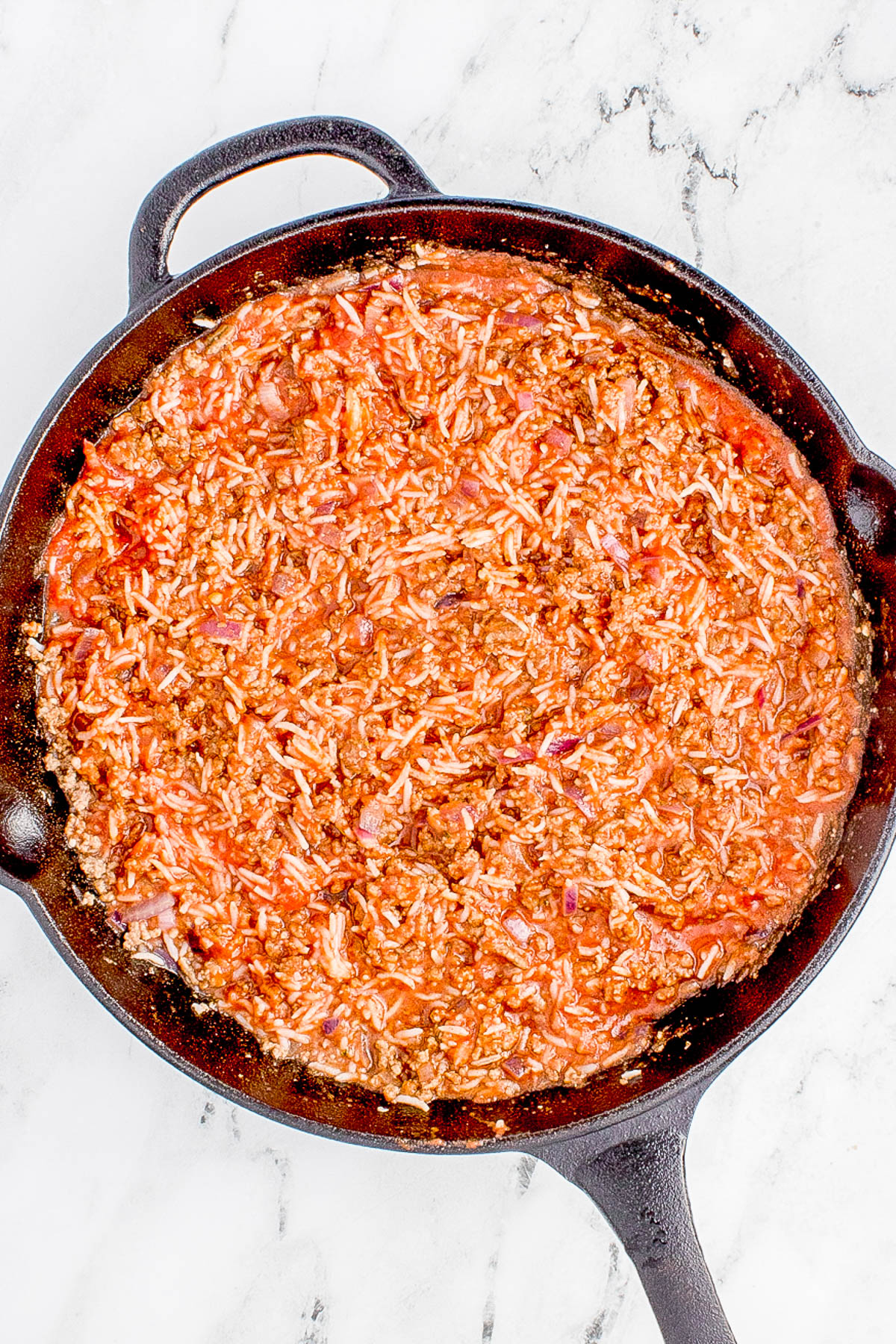 A skillet filled with a cooked mixture of ground meat, rice, and tomato sauce on a marble surface.