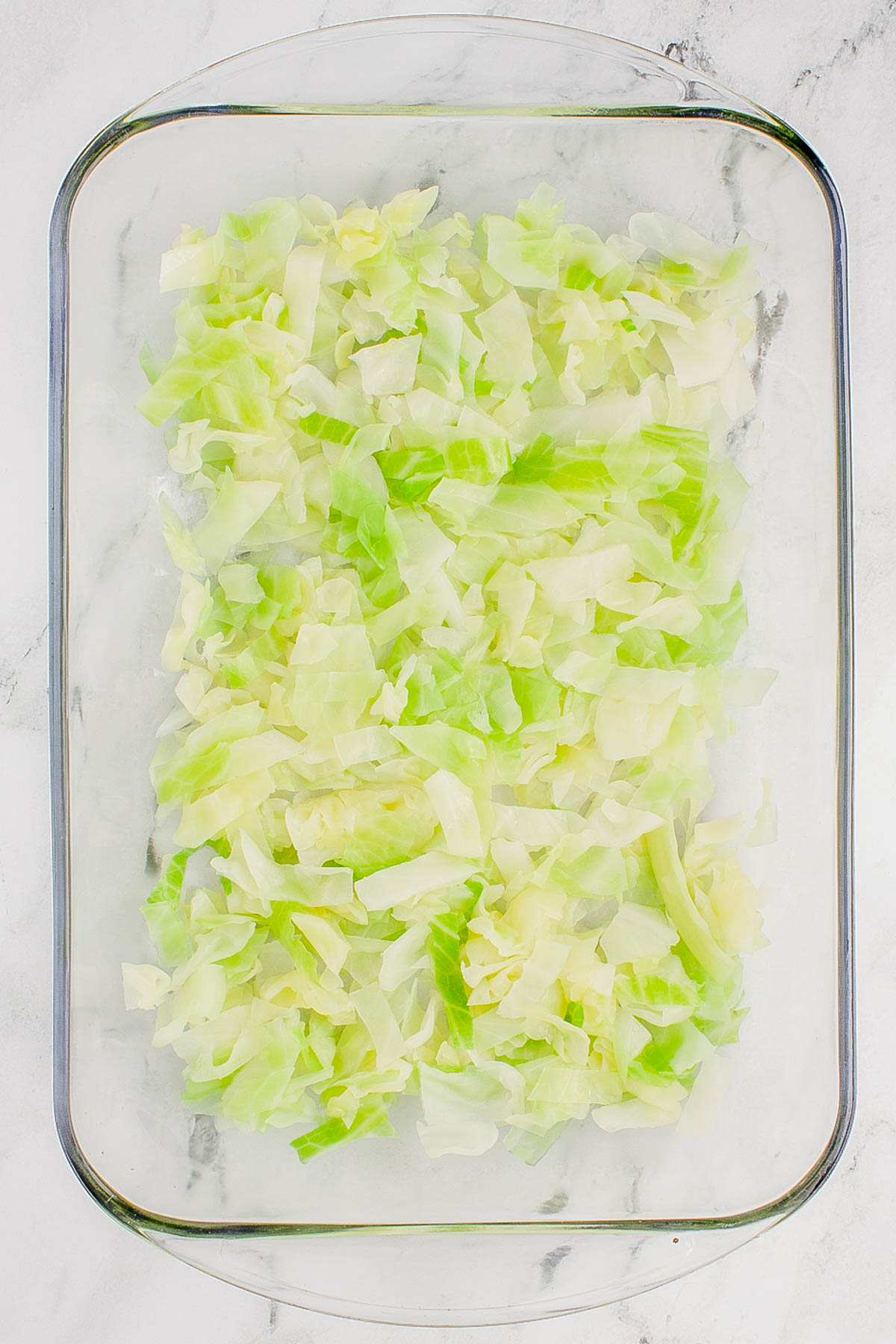 Chopped cabbage spread evenly in a transparent rectangular glass baking dish on a marble surface.