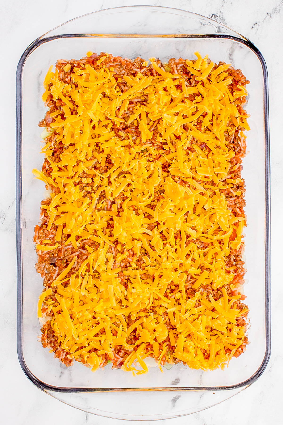 Glass baking dish with layered ground beef and shredded cheddar cheese on top.