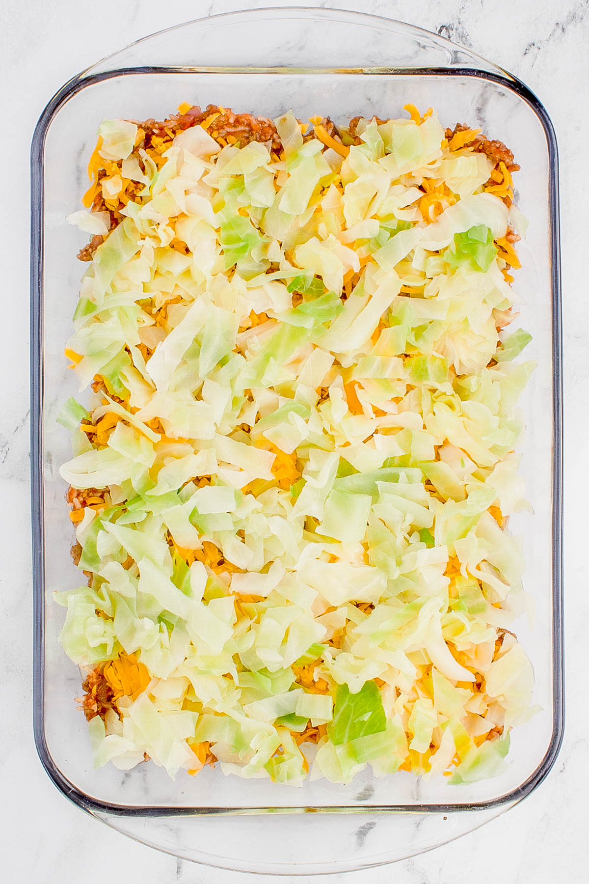 Glass baking dish with layers of chopped cabbage, melted cheese, and ground meat.