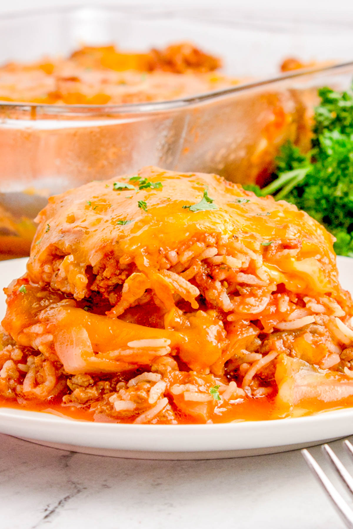 Close-up of a plate with a serving of cheesy beef and rice casserole, garnished with parsley. A glass baking dish with more casserole is visible in the background.