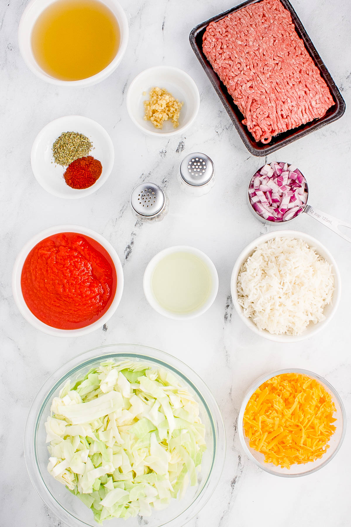 Ingredients laid out on a marble surface: ground meat, tomato sauce, rice, cabbage, cheese, onions, broth, spices, garlic, and oil in separate bowls.