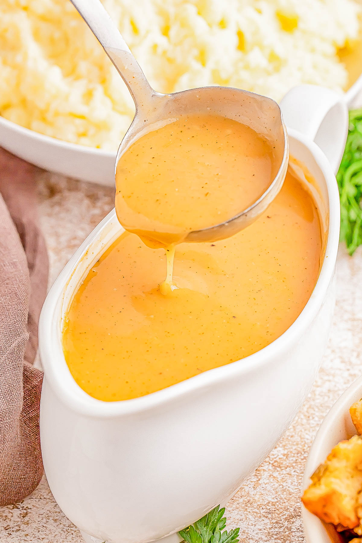 A ladle pouring creamy gravy into a white gravy boat, with a background of mashed potatoes.