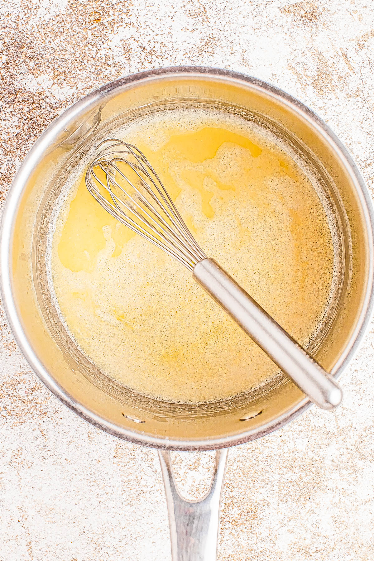 A whisk in a saucepan containing a yellow liquid mixture on a textured surface.