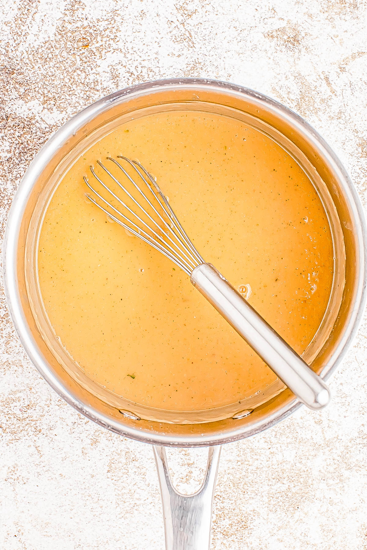 A whisk rests in a pot filled with smooth, amber-colored sauce set on a textured white background.