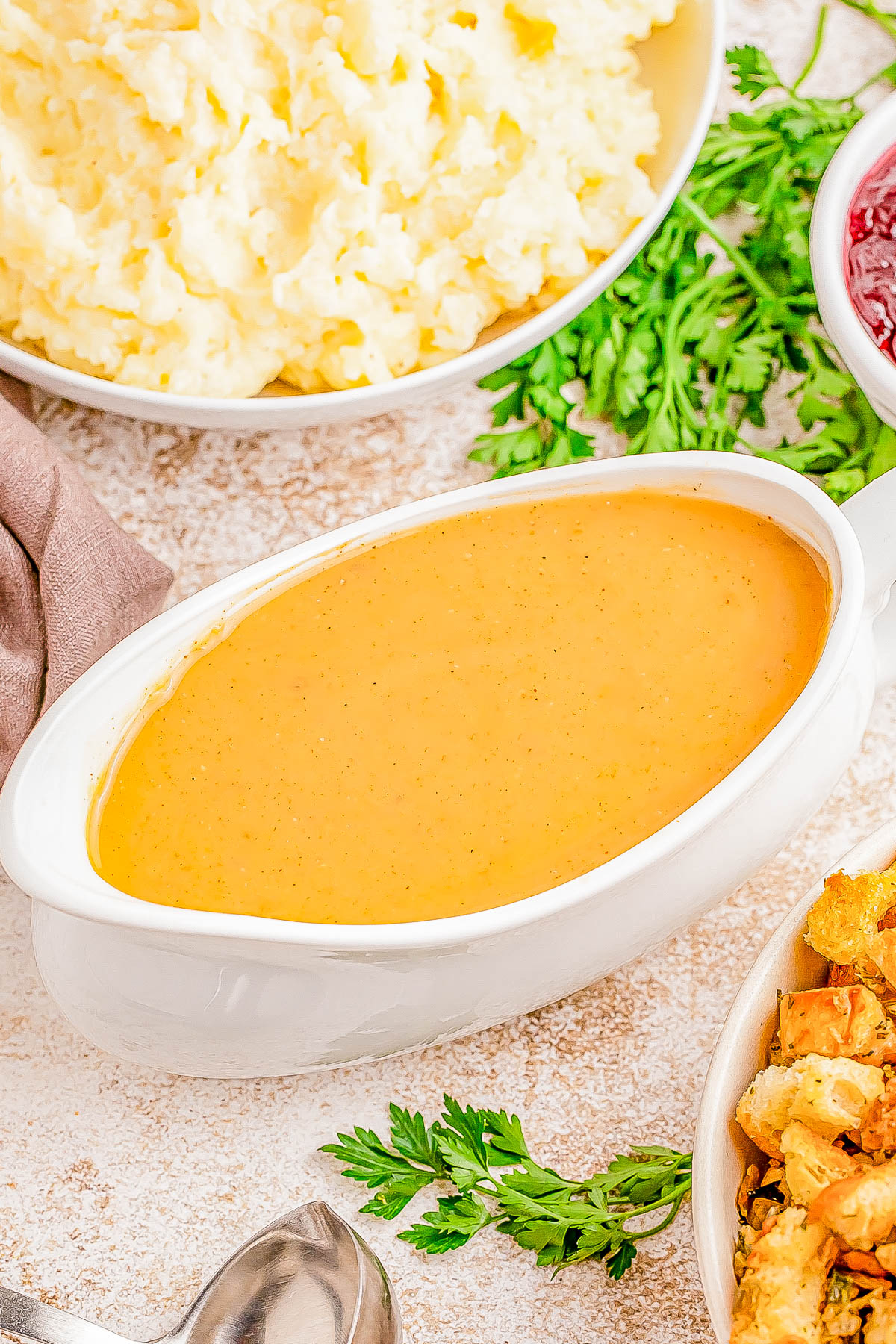 A white gravy boat filled with brown gravy, surrounded by mashed potatoes, stuffing, and fresh herbs on a light textured surface.